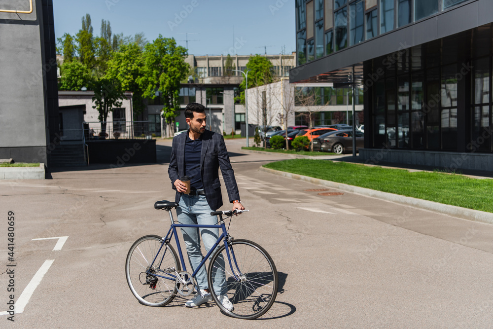 Young muslim manager in jacket holding coffee and bicycle on urban street