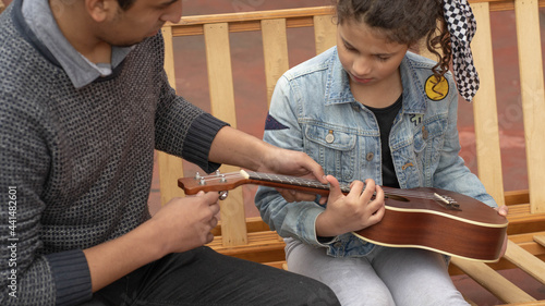 Young hispanic man teaches curly haired girl to tune ukulele outdoors music concept
