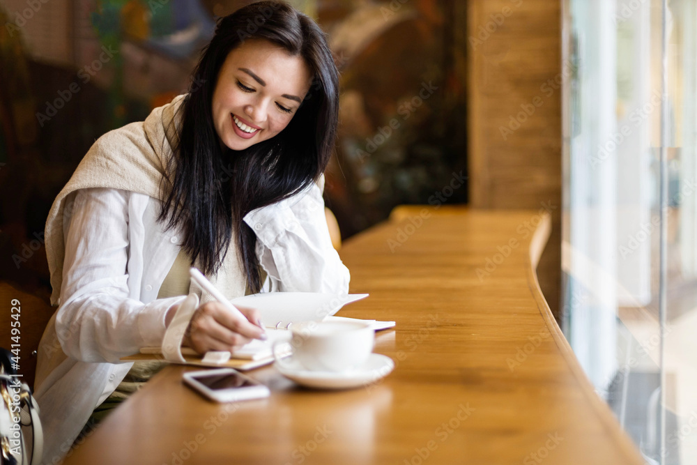 Happy Asian young woman sitting at cafe with coffee cup taking notes enjoying break Happy Asian young woman sitting at cafe with coffee cup taking notes enjoying break