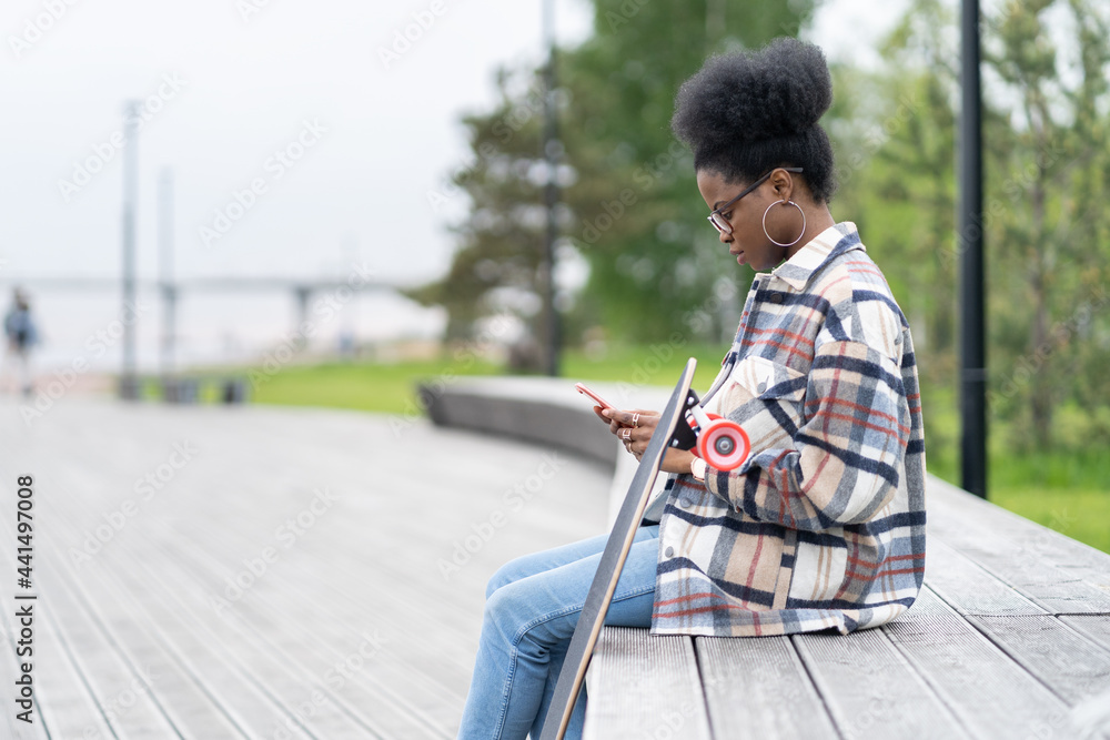 African woman skateboarder focused on reading sms messages in smartphone sit with longboard on bench in city park. Young trendy black female skater texting online outdoors in mobile phone application