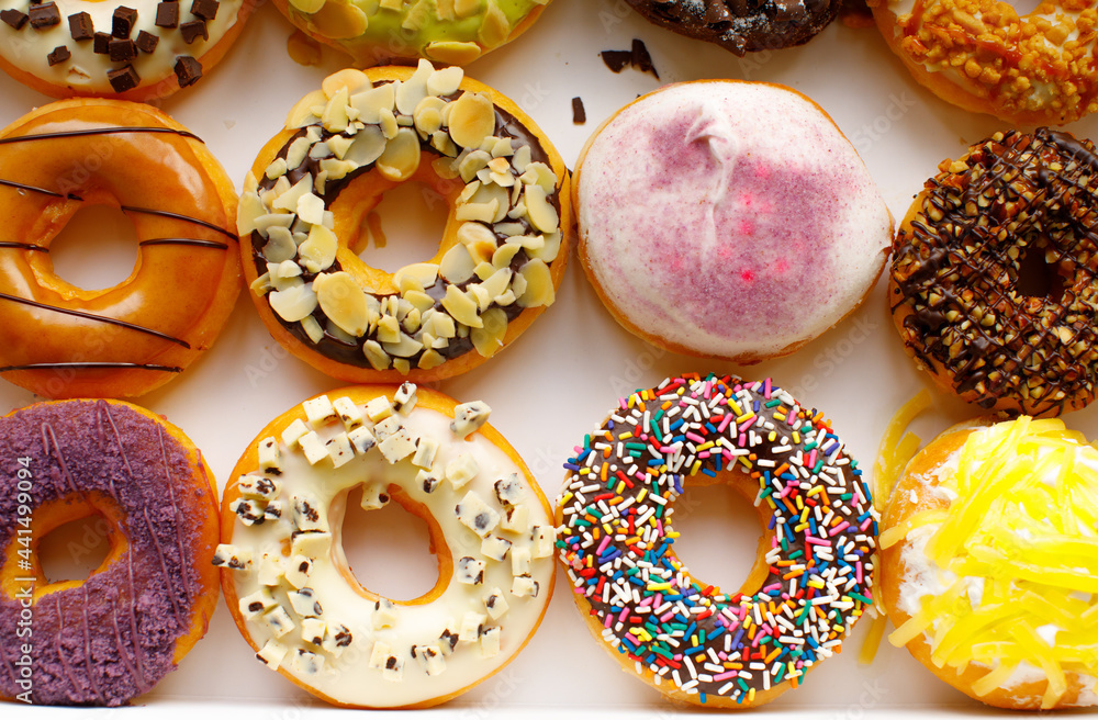 Top view flat lay of a colorful group of twelve delicious donuts in a white paper box on a wooden table. Collection of dozen donuts with glazes, sprinkles, almonds, peanuts, and chocolate chips Top view flat lay of a colorful group of twelve delicious donuts in a white paper box on a wooden table. Collection of dozen donuts with glazes, sprinkles, almonds, peanuts, and chocolate chips