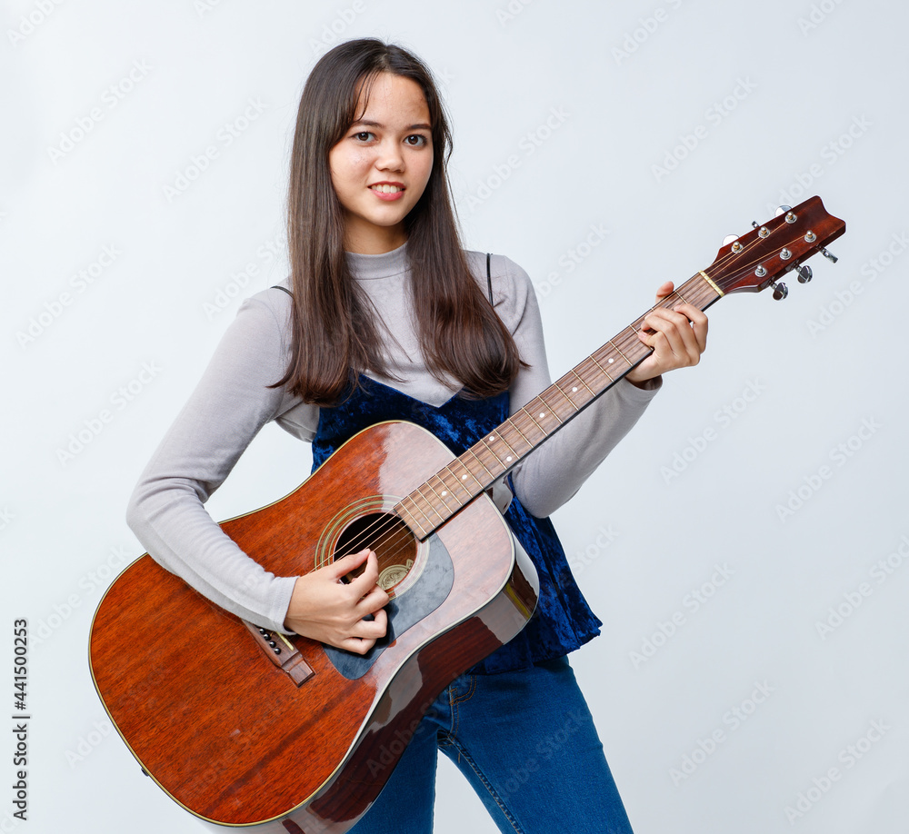 Portrait shot of a cute smiling Thai-Turkish teenager playing the acoustic guitar. Junior girl guitarist in casual clothes playing instrument while looking at the camera isolated in white background Portrait shot of a cute smiling Thai-Turkish teenager playing the acoustic guitar. Junior girl guitarist in casual clothes playing instrument while looking at the camera isolated in white background