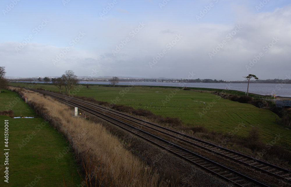 double track railway line in typical English farm land with a large river in the background with ablue sky and white clouds