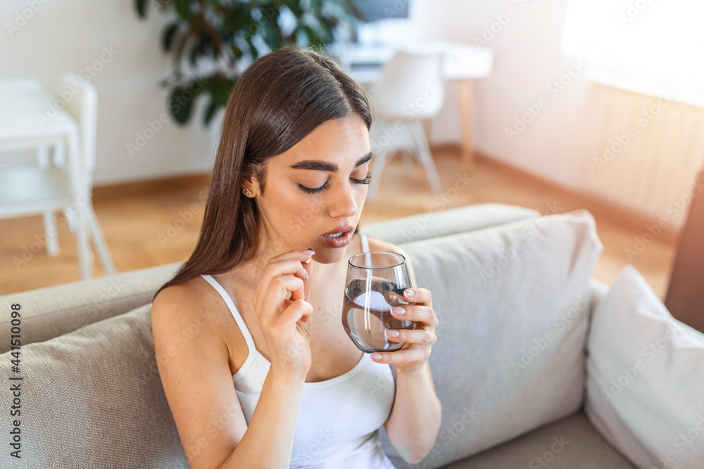 Concept of suffering from terrible pain. Beautiful, sad, upset, unhappy, troubled, weak woman with black hair, wearing casual clothes is sitting on a sofa and taking a pill Concept of suffering from terrible pain. Beautiful, sad, upset, unhappy, troubled, weak woman with black hair, wearing casual clothes is sitting on a sofa and taking a pill
