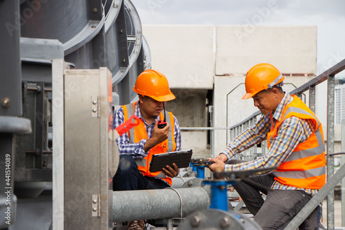 Asian engineer refrigeration plant  factory. male mechanic wearing helmet and Reflective Safety Vest uses laptop, tablet, equipment with sets of cooling towers conditioning systems background.