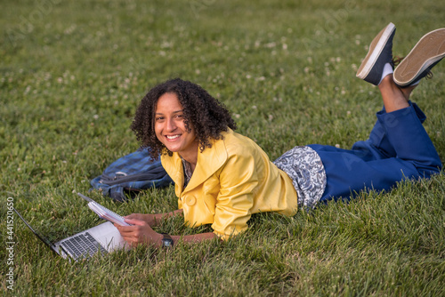 African American female student smiling at camera while laying on the lawn on campus; her laptop computer and notebook  in front of her