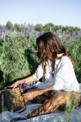 A fashionable Caucasian woman in a stylish white shirt feels free in a field with purple flowers in the sun. Lupin field in summer.The concept of a summer countryside. Close to outdoor recreation.
