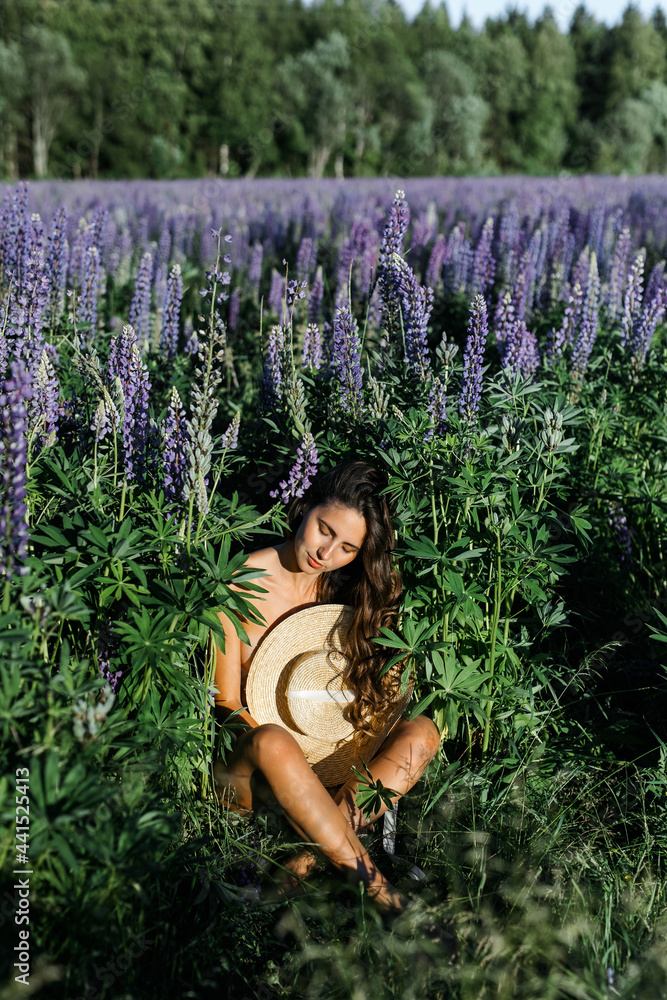portrait of a sexy girl in a straw hat. A fashionable Caucasian woman feels free in a field with purple flowers in the sun. Lupine field in summer. The concept of a summer countryside. Perfect body. portrait of a sexy girl in a straw hat. A fashionable Caucasian woman feels free in a field with purple flowers in the sun. Lupine field in summer. The concept of a summer countryside. Perfect body.
