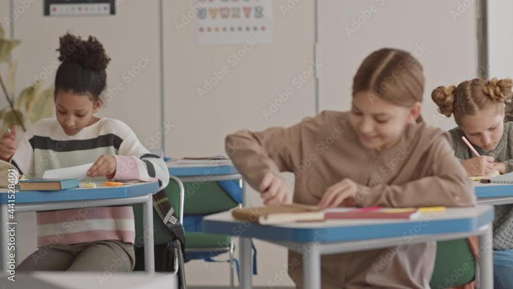 Medium shot of multiethnic schoolchildren sitting on by one in classroom, trying to cheat and exchange test papers while teacher is out of class Medium shot of multiethnic schoolchildren sitting on by one in classroom, trying to cheat and exchange test papers while teacher is out of class