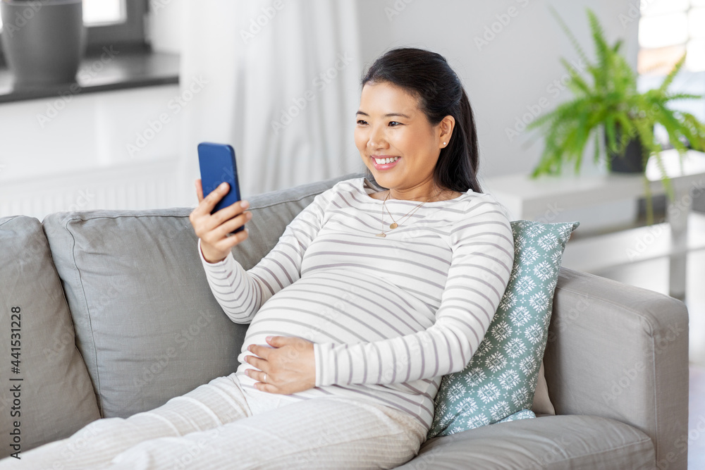pregnancy, rest, people and expectation concept - happy smiling pregnant asian woman with smartphone sitting on sofa at home pregnancy, rest, people and expectation concept - happy smiling pregnant asian woman with smartphone sitting on sofa at home