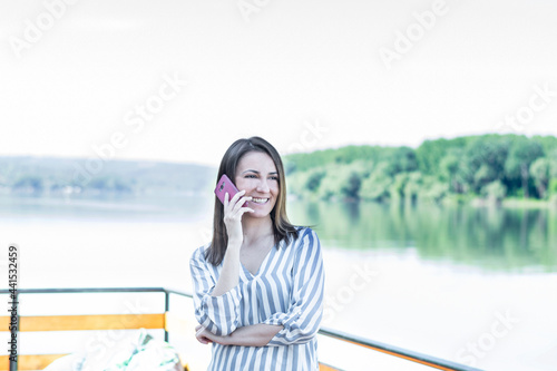 A young freelancer women works outside the office. She is on the phone at the cottage by the river. Forest and river in the background. A smiling girl is having a business conversation