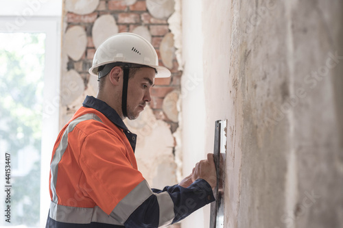 Tableau sur toile A man in a helmet and a work uniform aligns the walls with a spatula and putty