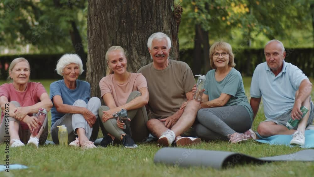 Portrait of happy young female fitness coach and group of cheerful senior men and women sitting on grass in park and looking together at camera Portrait of happy young female fitness coach and group of cheerful senior men and women sitting on grass in park and looking together at camera