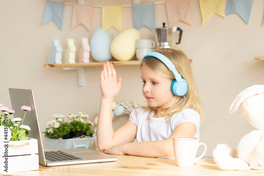 Smiling little blonde girl in headphones watches a video tutorial on the computer and raises her hand to answer. happy little kid in headphones has online class, using laptop at home, home learning Smiling little blonde girl in headphones watches a video tutorial on the computer and raises her hand to answer. happy little kid in headphones has online class, using laptop at home, home learning