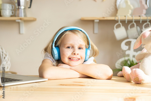 Little cute girl in headphones sits at the table and works at the computer. Looks at the camera and smiles. Happy children and homeschooling consuption photo