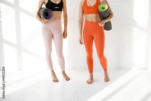 Two females in sportswear stand together with a yoga mats on a white wall background. Close-up on legs