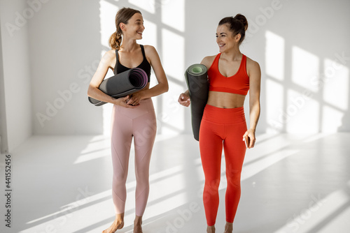 Two female friends in sportswear walk together with a yoga mats, talking and having fun during yoga break at gym