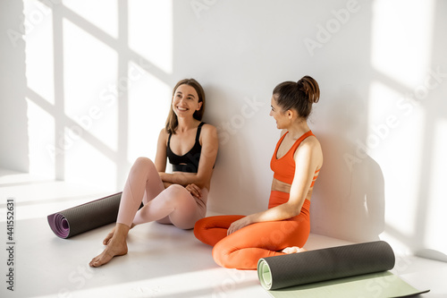 Two female friends in sportswear sitting together with yoga mats and having talk during a yoga training break at gym on the white background photo