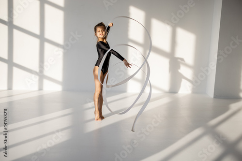Little girl practising rhythmic gymnastics with a gymnastic tape at white sunny dance room. Wide view with copy space and shadows on the background photo