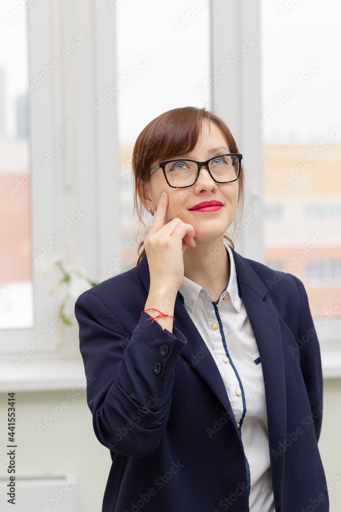 young stylish pretty business woman in a blue jacket and glasses by the window in the office at her workplace is satisfied with the work done young stylish pretty business woman in a blue jacket and glasses by the window in the office at her workplace is satisfied with the work done
