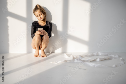 Foto Little girl sitting with smart phone during a break in rhythmic gymnastics class on a white wall background