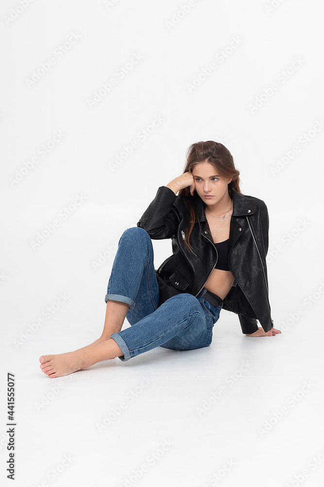 portrait of young caucasian attractive woman with long brown hair in blue jeans and black leather jacket on white background. skinny pretty lady posing at studio with bare feet