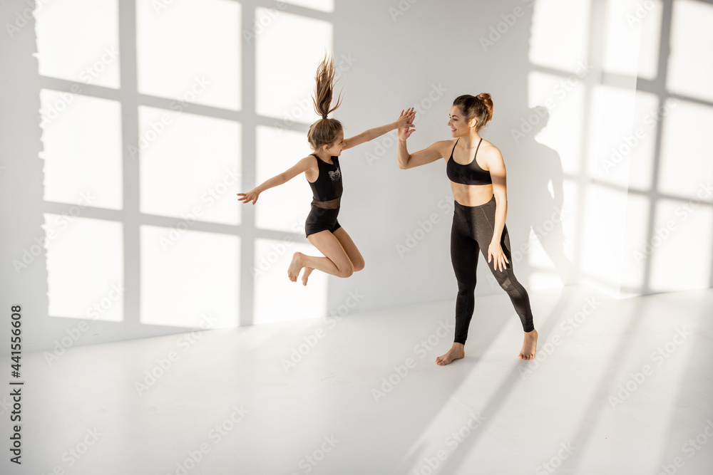 Naklejka premium Happy sports mom or trainer with a little girl during gymnastics training, jumping and having fun at white classroom