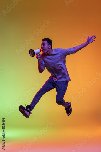 One Latina young man with megaphone, loudspeaker jumping high isolated on gradient yellow green background in neon light.