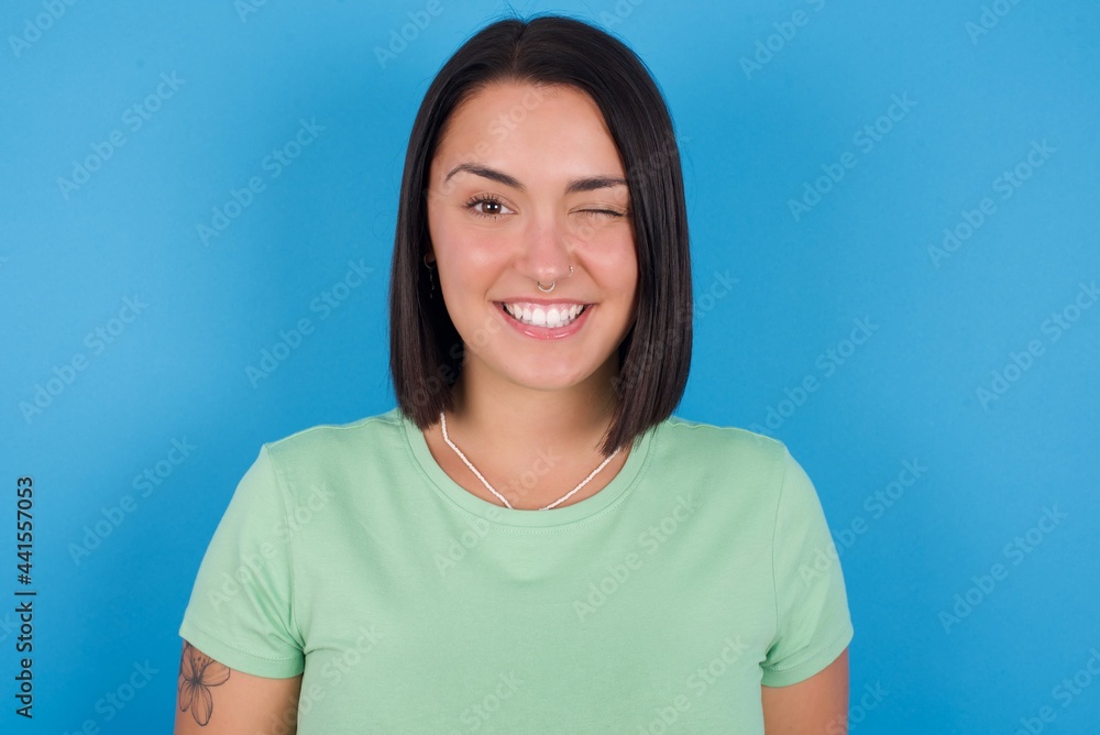 Coquettish young beautiful brunette girl with short hair standing against blue background smiling happily, blinking at camera in a playful manner, flirting with you. Coquettish young beautiful brunette girl with short hair standing against blue background smiling happily, blinking at camera in a playful manner, flirting with you.