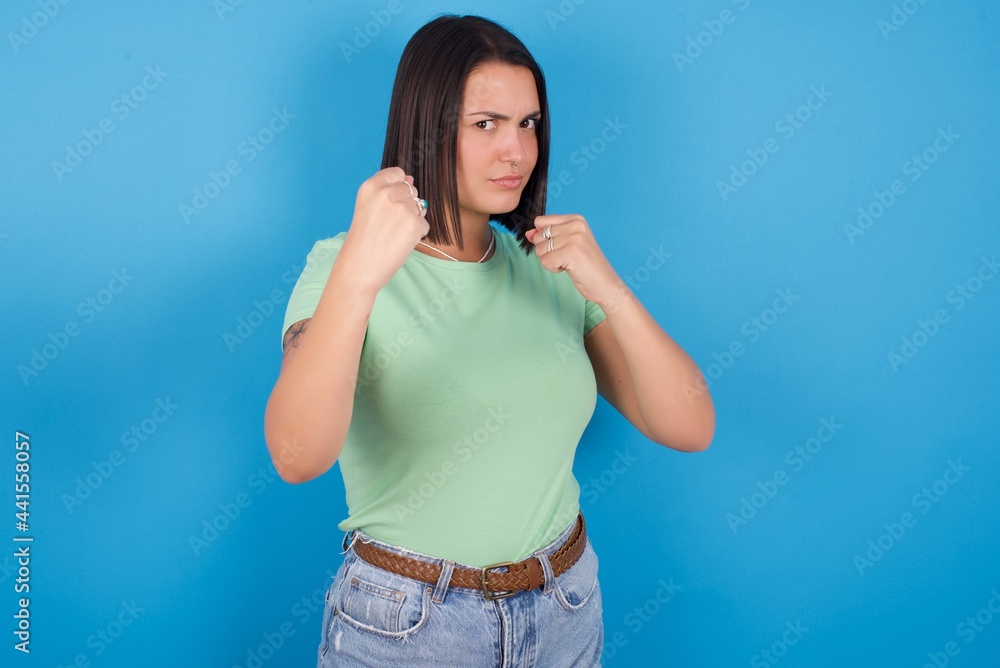 young beautiful brunette girl with short hair standing against blue background Ready to fight with fist defense gesture, angry and upset face, afraid of problem. young beautiful brunette girl with short hair standing against blue background Ready to fight with fist defense gesture, angry and upset face, afraid of problem.