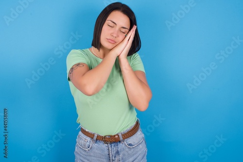 young beautiful brunette girl with short hair standing against blue background sleeping tired dreaming and posing with hands together while smiling with closed eyes.