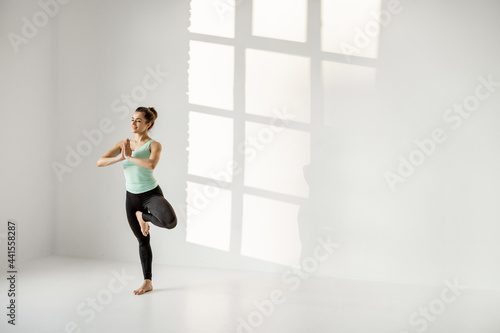 Athletic woman standing in meditation pose, practising yoga at white sunny classroom alone. Wide view with copy space © rh2010