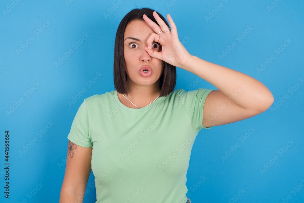 young beautiful brunette girl with short hair standing against blue background doing ok gesture shocked with surprised face, eye looking through fingers. Unbelieving expression. young beautiful brunette girl with short hair standing against blue background doing ok gesture shocked with surprised face, eye looking through fingers. Unbelieving expression.