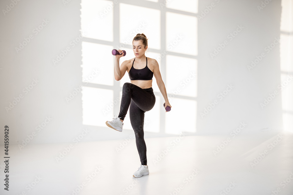 Beautiful and confident athletic woman doing cardio with dumbbells at white room. Training fitness or aerobics alone