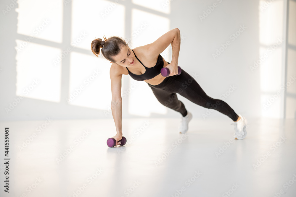 Fototapeta premium Beautiful and confident athletic woman doing cardio with dumbbells at white room. Training fitness or aerobics alone