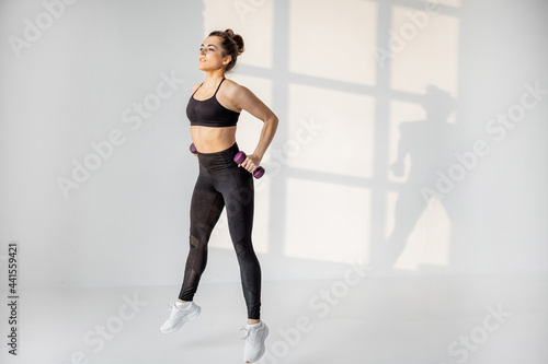 Beautiful and confident athletic woman doing cardio with dumbbells at white room. Training fitness or aerobics alone