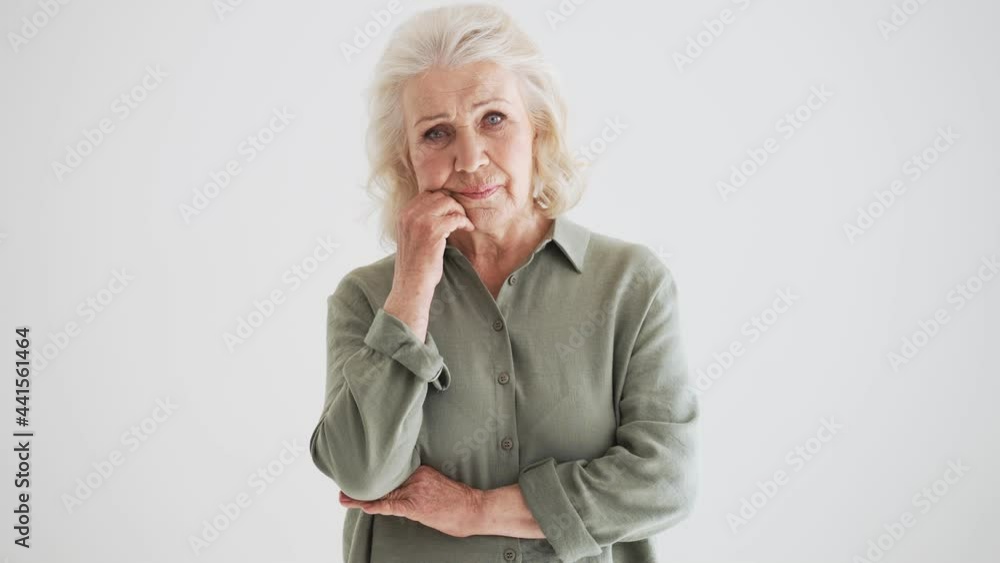 A bored elder woman is looking to the camera standing isolated over white wall in the studio A bored elder woman is looking to the camera standing isolated over white wall in the studio