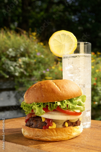 A close-up of lemonaid and a hamburger topped with melted cheese, bacon, onion, tomato, lettuce, ketchup and mustard on wooden tabletop outdoors with flowers in the background photo