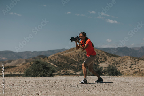 Woman photographer in action, taking photos in Tabernas Desert, Spain, in a sunny day, with an orange color t-shirt.