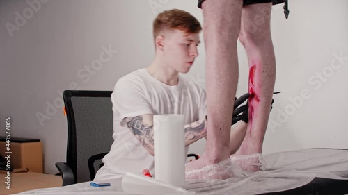 Young ginger man tattoo artist adding finishing touches to a sketch on the leg on his client with a pen over the red marker
