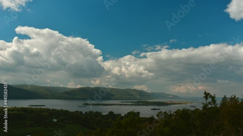 Wide river flowing between high hills filmed in time lapse. The Volga river near the Zhigulevsky mountains. Summer landscape. Green mountains under the blue sky. Steamers move along the river.