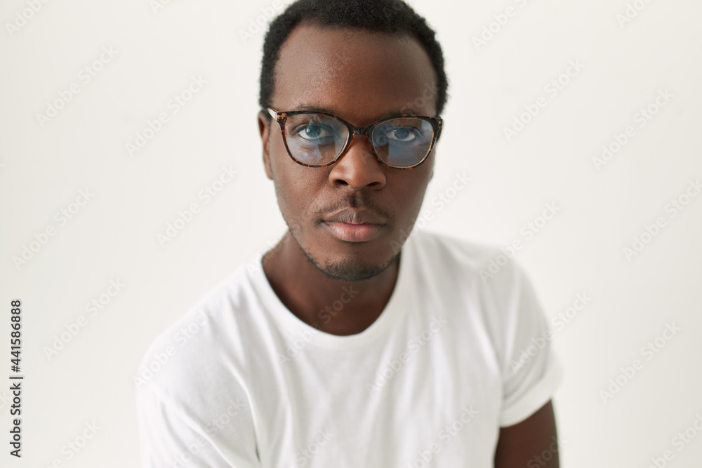 Close up image of handsome young dark skinned man with trimmed stubble posing isolated wearing stylish glasses and white tee looking at camera with confident facial expression. Eyewear and vision Close up image of handsome young dark skinned man with trimmed stubble posing isolated wearing stylish glasses and white tee looking at camera with confident facial expression. Eyewear and vision