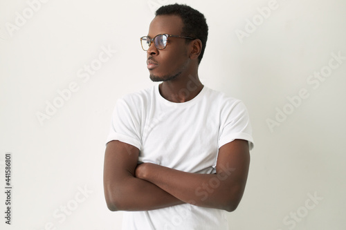 Isolated shot of serious stubborn young African male wearing blank white t-shirt and eyeglasses crossing arms on his chest, looking away, expressing disagreement, ignoring you, having grumpy look