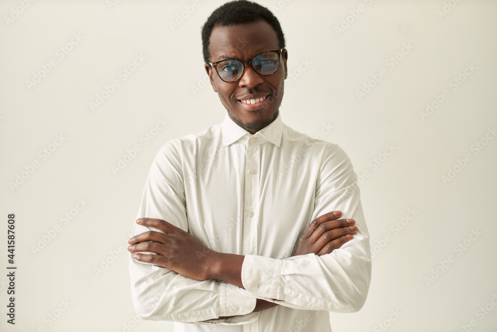 Studio image of attractive unshaven young dark skinned manager wearing formal shirt and eyeglasses crossing arms on his chest and smiling feeling confident and proud of himself. Body language Studio image of attractive unshaven young dark skinned manager wearing formal shirt and eyeglasses crossing arms on his chest and smiling feeling confident and proud of himself. Body language