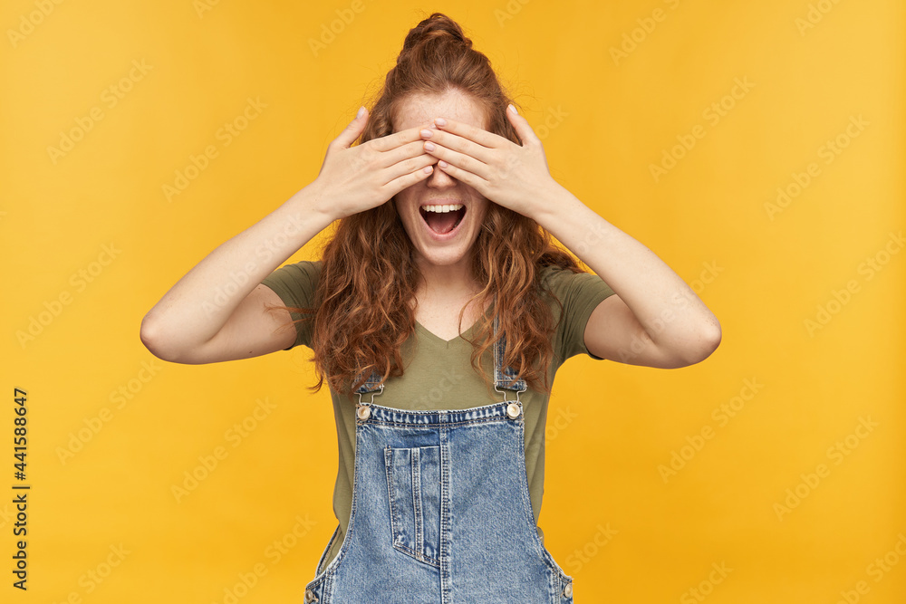 portrait of funny young ginger female wears blue overalls and green t-shirt, smiles broadly and close her eyes with her palms waiting for surprise. isolated over yellow background. portrait of funny young ginger female wears blue overalls and green t-shirt, smiles broadly and close her eyes with her palms waiting for surprise. isolated over yellow background.