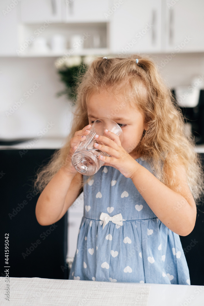 Toddler child drinking water at home from the glass on the kitchen. Kid girl is thirsty. Offer crystal still mineral water for body refreshment Toddler child drinking water at home from the glass on the kitchen. Kid girl is thirsty. Offer crystal still mineral water for body refreshment