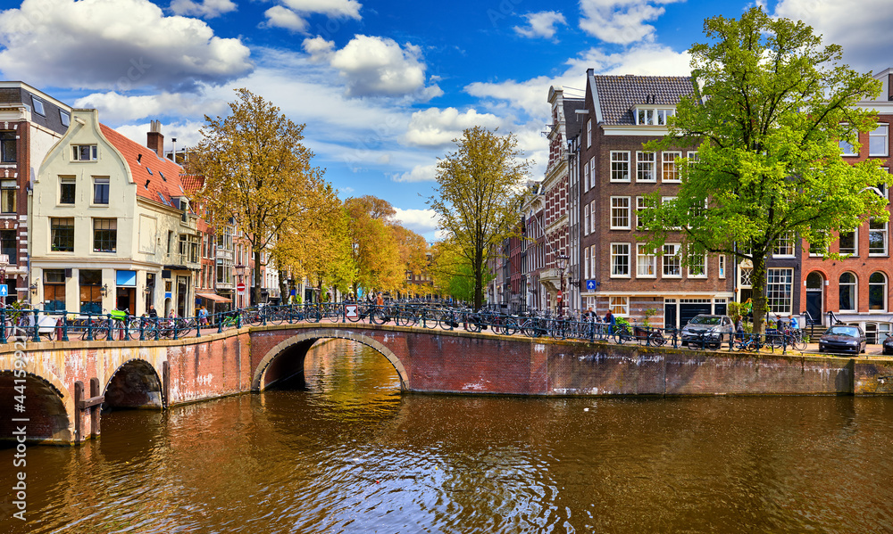 Netherlands Amsterdam. View at river Amstel with traditional houses at bank and boats at water canal. Scenic cityscape. Spring day with blue sky and white clouds.