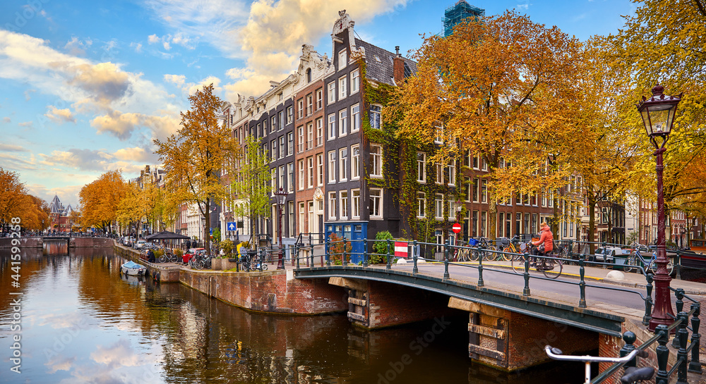 Netherlands Amsterdam. View at river Amstel with traditional houses at bank and boats at water canal. Scenic cityscape. Spring day with blue sky and white clouds.
