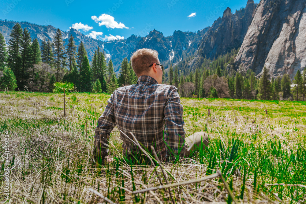 A young man is sitting on a green lawn with an amazing view of the mountains in Yosemite National ​Park