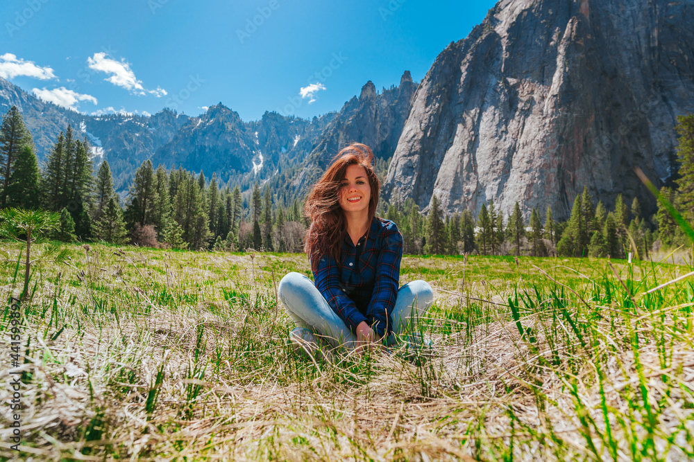 A young woman in jeans sits on the grass with an amazing view of the mountains in Yosemite National Park